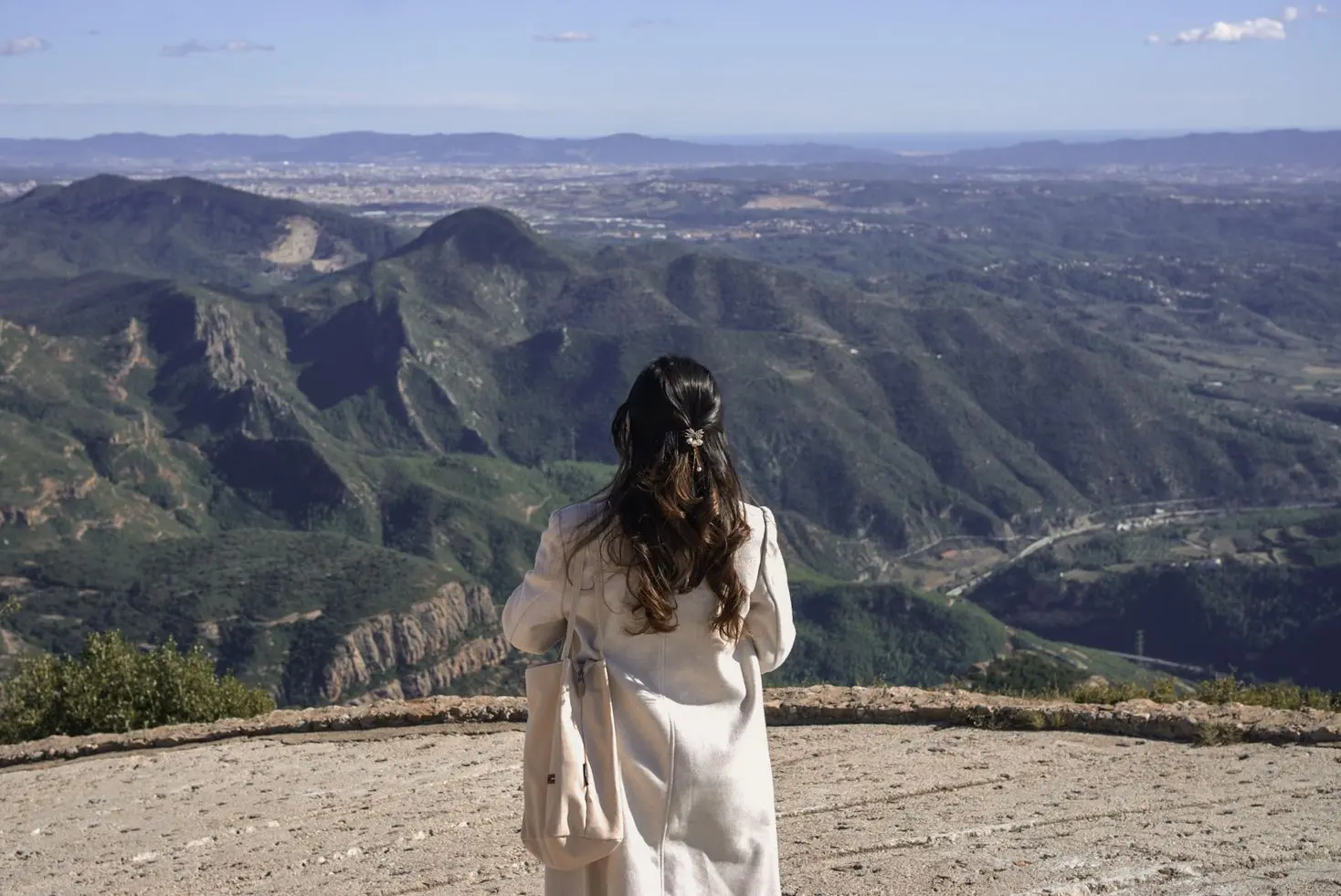 Woman from behind on a scenic mountain overlook, viewing a vast landscape with green hills, a distant city, and the sea.