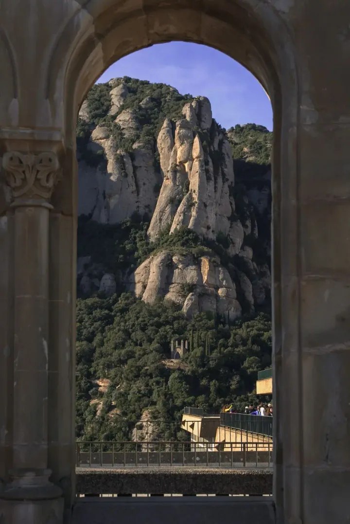 Rugged Montserrat mountains and lush trees framed by a stone archway. A distant chapel and a viewing platform are visible.